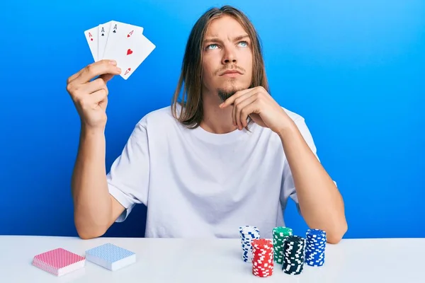 英俊的高加索男人 留着长发打扑克 拿着扑克牌 严肃的脸 手托着下巴思考着问题 沉思着困惑的想法 — 图库照片
