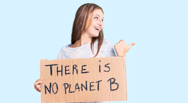 Young beautiful blonde woman holding there is no planet b banner pointing thumb up to the side smiling happy with open mouth
