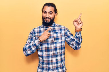 Gündelik kıyafetler giyen genç Arap adam gülümseyip kameraya bakıyor ve iki eliyle ve parmaklarıyla kenarı işaret ediyor.. 