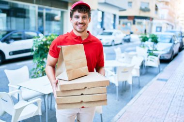 Beyaz tenli genç teslimatçı elinde mutlu bir gülümsemeyle elinde pizza kartonuyla şehirde yürüyor..