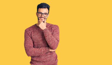 Kışlık süveter ve gözlük takan yakışıklı genç adam kameraya kendinden emin bir şekilde bakıyor kollarını kavuşturup, çenesini kaldırarak gülümsüyor. Olumlu düşünmek. 