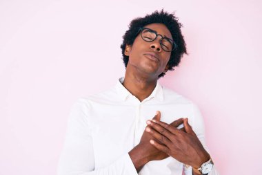 Afro saçlı, iş gömleği giyen, gözlüklü, göğsünde elleri göğsünde gülümseyen, yüzünde minnet ifadesi olan yakışıklı Afro-Amerikan erkeği. sağlık kavramı. 