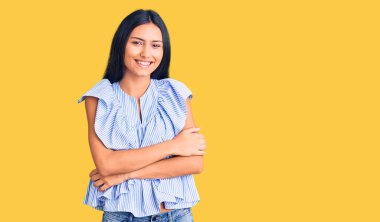 Gündelik kıyafetler giyen, kollarını kavuşturup kameraya gülümseyen genç, güzel bir Latin kız. pozitif kişi. 