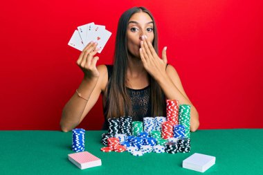Masada oturmuş poker oynayan, ağzı elle kapatan, şok olmuş ve hata yapmaktan korkan genç İspanyol kadın. şaşırmış ifade 
