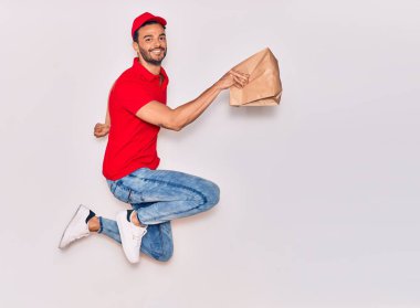 Mutlu gülümseyen üniformalı yakışıklı İspanyol teslimatçı. Yüzünde tebessümle, elinde torbayla, izole edilmiş beyaz arka planda paket yiyeceklerle zıplıyorsun..