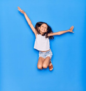 Sevimli İspanyol kız günlük kıyafetler giyip mutlu gülümsüyor. İzole edilmiş mavi arka planda yüzünde gülümsemeyle zıplıyor