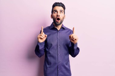 Genç, sakallı, sıradan gömlekli yakışıklı adam şaşırmış ve şaşırmış bir şekilde yukarı bakıyor ve parmaklarıyla işaret ediyor ve kollarını kaldırıyor.. 