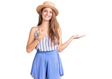 Sarışın, güzel bir kadın, yaz şapkası takıyor, avuç içini gösteriyor ve baş parmaklarıyla iyi bir hareket yapıyor, mutlu ve neşeli gülümsüyor. 