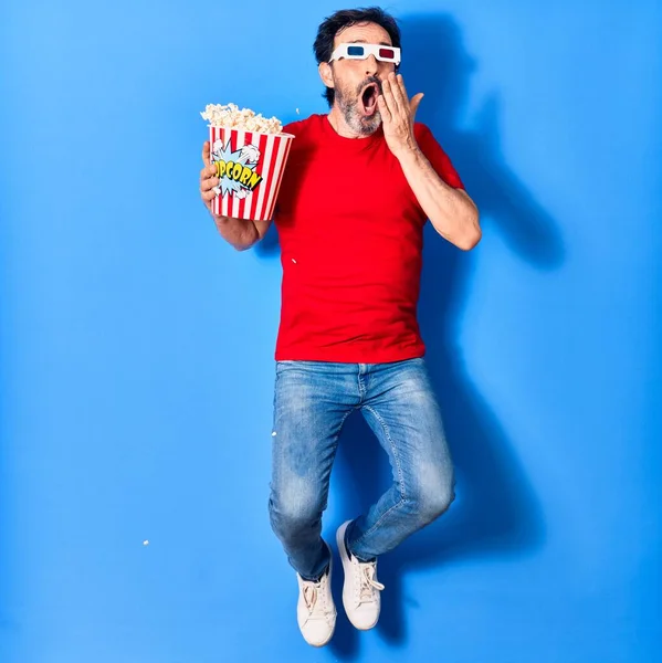 Middle age handsome hispanic man suprised with open mouth watching movie using 3d glasses. Holding bucket of popcorn jumping over isolated blue background