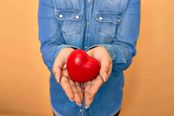 Woman holding red heart with two hands standing over isolated yellow background