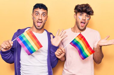 Genç eşcinsel çift ellerinde gökkuşağı lgbtq bayrakları ile zaferi kutluyorlar mutlu bir gülümsemeyle ve kazanan bir ifadeyle. 