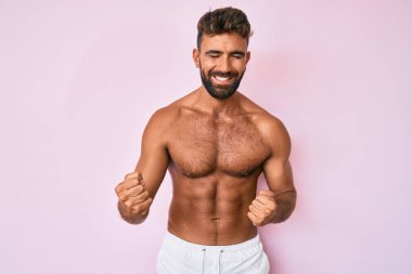 Genç İspanyol adam üstsüz, çok mutlu ve heyecanlı bir şekilde kollarını kaldırıp, gülümseyerek ve başarı için bağırarak kazanan jestler yapıyor. kutlama konsepti. 