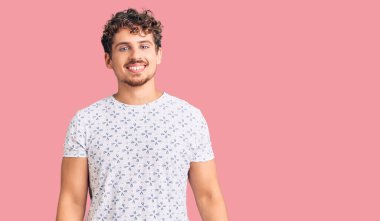 Kıvırcık saçlı, sıradan giyinen, pozitif ve mutlu görünen, kendine güvenen gülümseyen, dişlerini gösteren genç ve yakışıklı bir adam. 