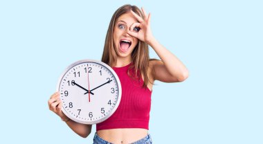 Genç, güzel, sarışın bir kadın, kocaman bir saat tutuyor, gülümsüyor. 