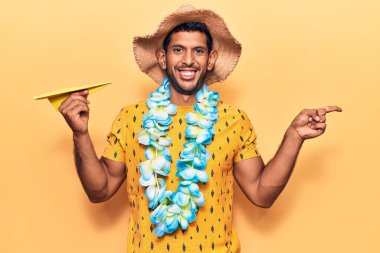 Yaz şapkası takan genç bir latin ve elinde kağıt uçakla gülümseyen Hawaii zambağı tutan mutlu bir adam. 
