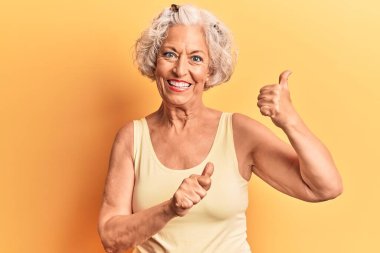 Kıdemli, gri saçlı, günlük kıyafetler giyen, arka tarafı işaret eden, el ve baş parmakları havada, kendine güvenen bir kadın. 