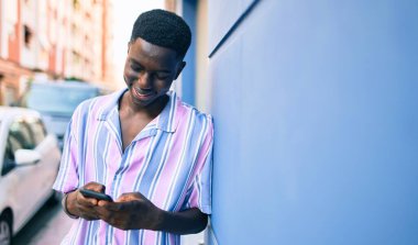 Afro-Amerikalı genç adam mutlu gülümsüyor. Şehrin caddesinde duvara yaslanmış akıllı telefon kullanıyor..