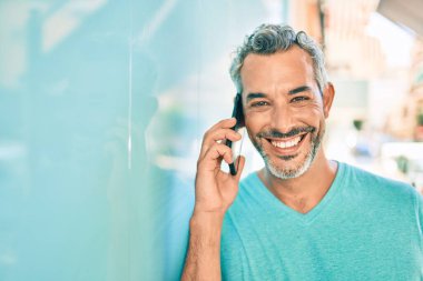 Orta yaşlı, kır saçlı, akıllı telefondan konuşan ve şehrin duvarına yaslanan bir adam..