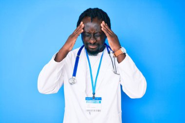 Doktor steteskop ve kimlik kartı takan, başı ağrıyan, stresli ve çaresiz bir şekilde acı çeken, saçları örülmüş genç Afrikalı Amerikalı bir adam. Eller başının üstünde.. 