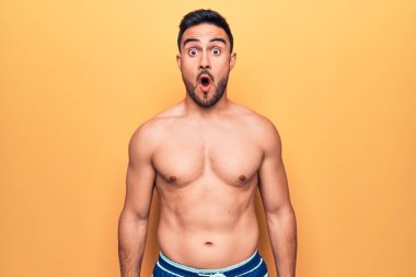 Sakallı, kolsuz tişörtlü, sarı arka planda duran yakışıklı bir genç. Şaşırmış ve ağzı açık kalmış.