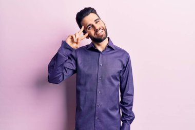 Sakallı, sıradan gömlekli, barış sembolü yapan, yüzünde parmakları olan, neşeli bir gülümsemeyle zaferi gösteren genç yakışıklı bir adam. 