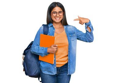 Sırt çantası takan, elinde kitaplarla gülümseyen ve parmaklarıyla ve ağzıyla işaret eden genç Latin kız. diş sağlığı kavramı. 