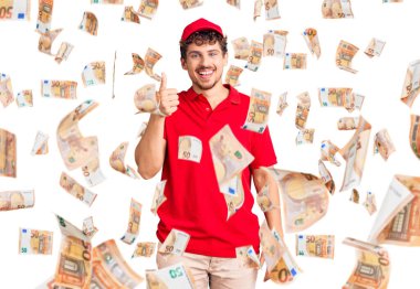 Kıvırcık saçlı, teslimat üniforması giyen, mutlu ve pozitif gülümseyen, başparmağıyla mükemmel ve onay işareti yapan genç yakışıklı bir adam.