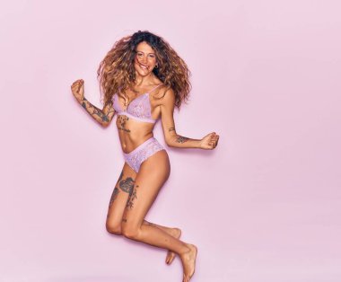 Dövmeli, iç çamaşırlı, mutlu gülümseyen genç bir kadın. Yüzünde tebessümle zıplıyor, pembe arka planda yumruklar havada kutluyor.