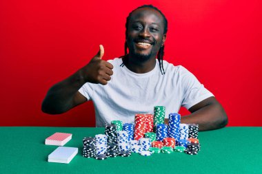 Poker fişleri ve kartlarıyla masada oturan yakışıklı genç adam mutlu ve olumlu gülümsüyor, başparmağıyla mükemmel ve onay işareti yapıyor. 
