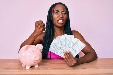 Domuz kumbarası ve dolarlarla masada oturan Afrikalı Amerikalı kadın öfkeyle bağırıyor, öfkeyle bağırıyor ve el kaldırıyor. 