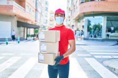 Üniforma giyen ve Coronavirus koruması takan genç bir teslimatçı. Sağlık maskesi. Kasaba sokaklarında karton paketler tutuyor..
