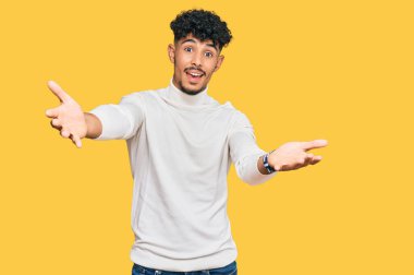 Genç Arap adam, sıradan bir kazak giyip kameraya bakıyor ve sarılmak için kollarını açmış gülümsüyor. Mutluluğu kucaklayan neşeli bir ifade.. 