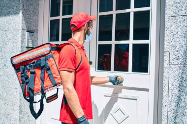 Beyaz teslimatçı kırmızı üniforma ve teslimat çantası ve koronavirüs güvenlik maskesi takıyor.