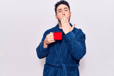 Bornoz giymiş genç İspanyol bir adam elinde kahveyle ağzını eliyle kapatıyor, şaşırmış ve hata yapmaktan korkuyor. şaşırmış ifade 