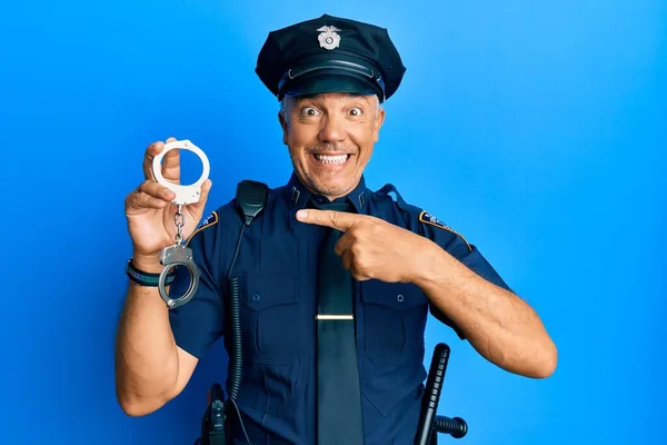 Handsome middle age mature man wearing police uniform holding metal handcuffs smiling happy pointing with hand and finger