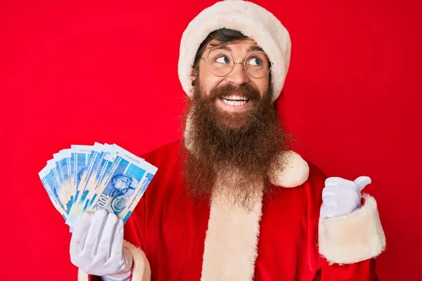 Handsome young red head man with long beard wearing santa claus costume holding african rand pointing thumb up to the side smiling happy with open mouth