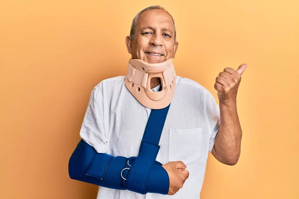 Handsome mature senior man wearing cervical collar and arm on sling pointing to the back behind with hand and thumbs up, smiling confident