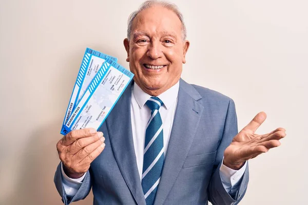 Handsome grey-haired man wearing suit holding airline boarding pass over white background celebrating achievement with happy smile and winner expression with raised hand