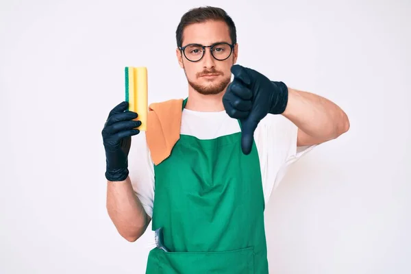 Young handsome man wearing apron holding scourer with angry face, negative sign showing dislike with thumbs down, rejection concept