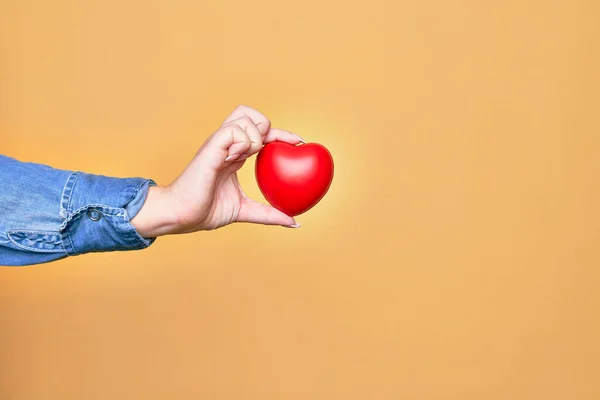 Mão Caucasiano Jovem Segurando Coração Vermelho Sobre Fundo Amarelo Isolado — Fotografia de Stock