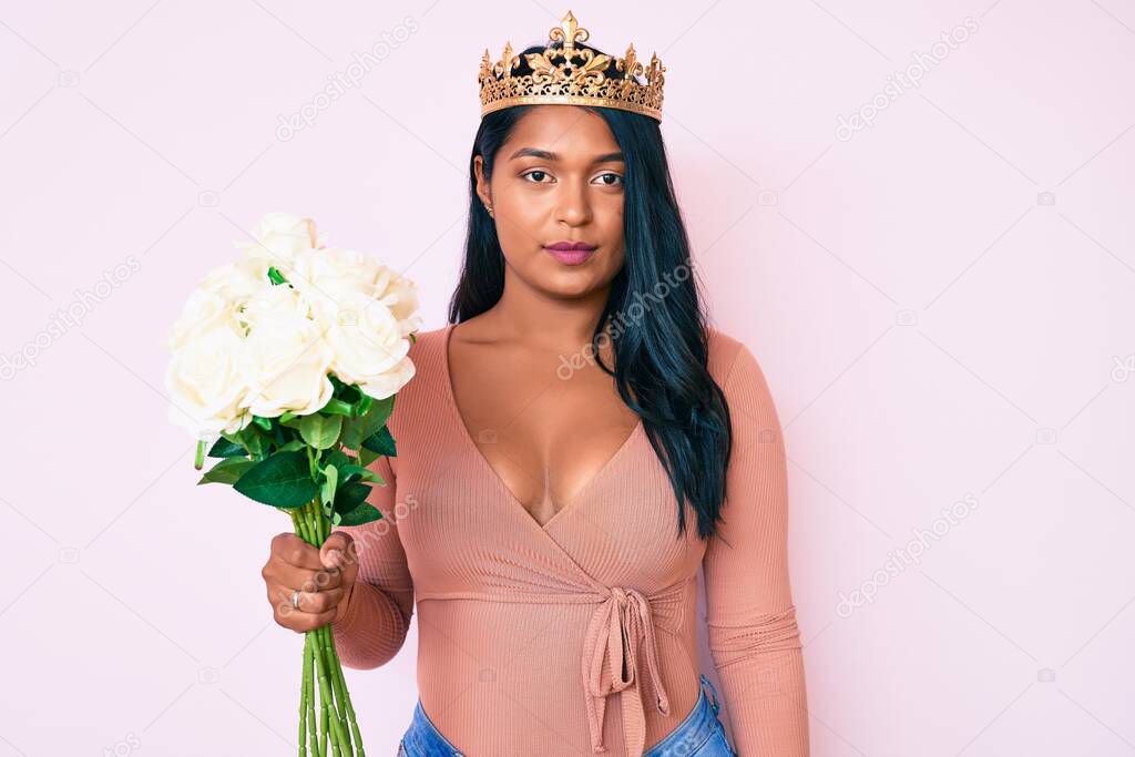 Beautiful latin young woman with long hair wearing princess crown and holding flowers thinking attitude and sober expression looking self confident 