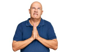 Orta yaşlı kel bir adam gündelik giysiler giyip elleri bağlı yalvarıyor ve dua ediyor. Yüzünde umut ifadesi var. Çok duygusal ve endişeli. Yalvarıyor. 