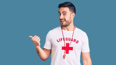 Cankurtaran üniforması giyen, gülümseyen, gülümseyen ve başparmağıyla yan tarafı gösteren genç yakışıklı bir adam.. 