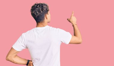 Genç ve yakışıklı bir adam, günlük beyaz tişört giymiş, tersten poz veriyor ve parmağıyla öne bakıyor. 