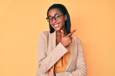 Genç Afro-Amerikan kadın iş kıyafetleri giymiş, yüzünde kocaman bir gülümseme var, eli ve parmağı kameraya bakıyor.. 
