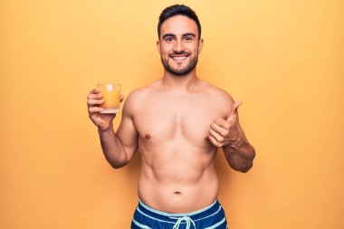 Tatilde sakallı, mayo giyen, portakal suyu içen mutlu ve pozitif gülümseyen, baş parmağını kaldıran, mükemmel ve onay işareti yapan genç yakışıklı bir adam.