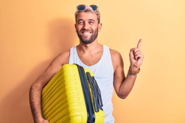 Genç, sarışın, tatilde olan güneş gözlüklü bir turist. Elinde uçak çantası var. Gülümsüyor, mutlu bir şekilde elini ve parmağını yana uzatıyordu.