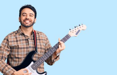 Genç İspanyol adam elektro gitar çalıyor. Pozitif ve mutlu görünüyor. Kendine güvenen bir gülümsemeyle ayakta duruyor ve dişlerini gösteriyor. 