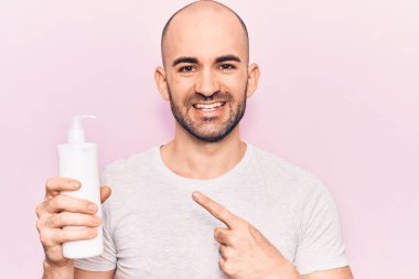 Genç yakışıklı kel adam elinde krem losyon şişesiyle gülümsüyor. 
