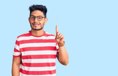 Yakışıklı Latin Amerikalı gündelik kıyafetler ve gözlük takan genç adam bir numarayı gösterip işaret ederken kendinden emin ve mutlu bir şekilde gülümsüyor.. 